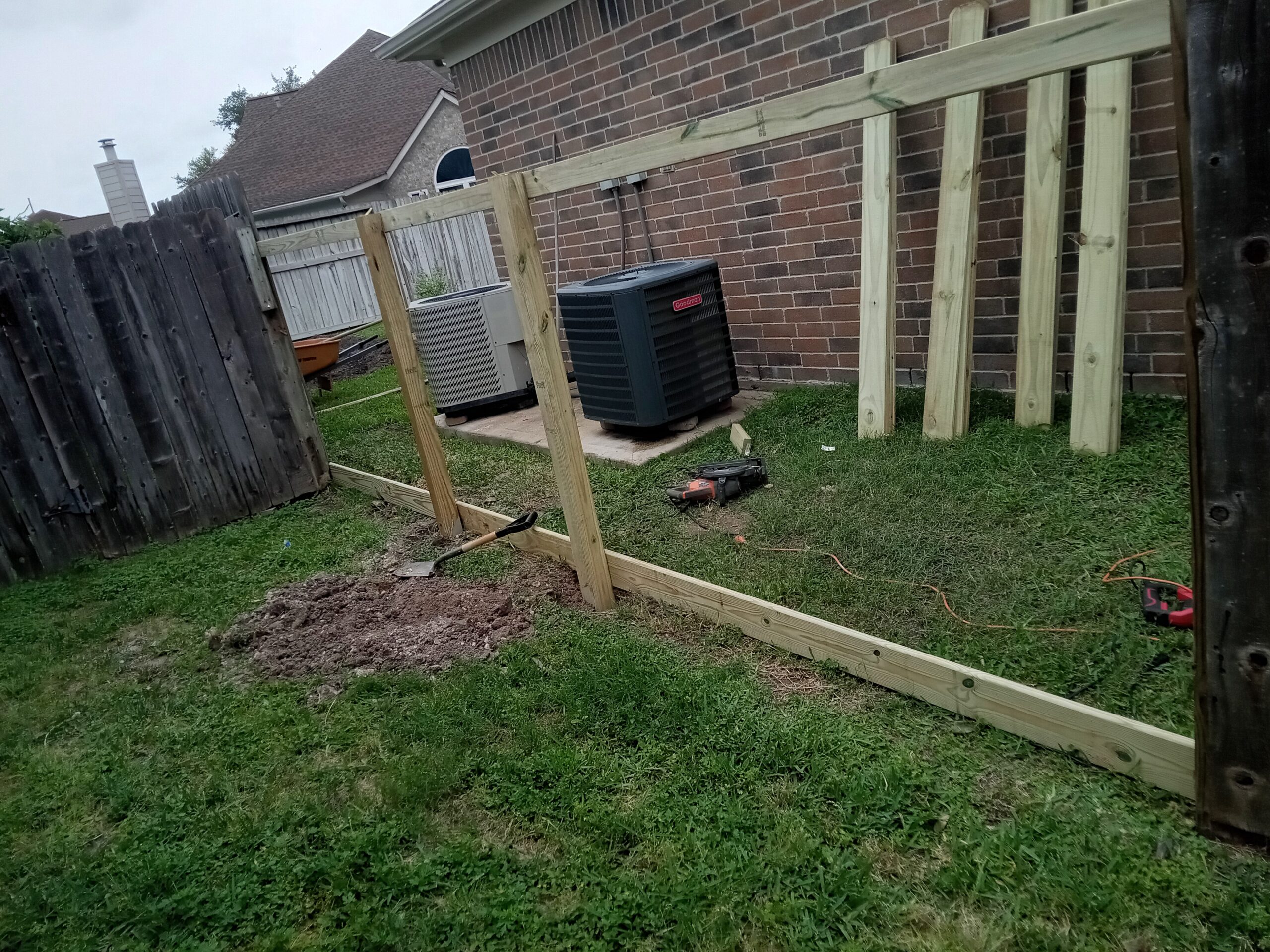 An unfinished wooden fence repair in progress in a Houston backyard. Two new fence posts have been erected and are supported by temporary braces, standing beside a section of old, weathered fence that remains upright. Excavated soil is visible at the base of the new posts.